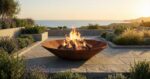 Low-profile Corten steel fire pit Diamond Octagon on a coastal terrace at sunset, with sea view emphasizing the warm rust patina.