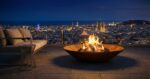 Luxury Diamond Octagon outdoor fire pit on a rooftop terrace at night, with illuminated city skyline in the background.